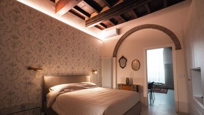 Quarto com cama, mesa de cabeceira, cômoda, espelho e uma porta arqueada que dá para outro cômodo.