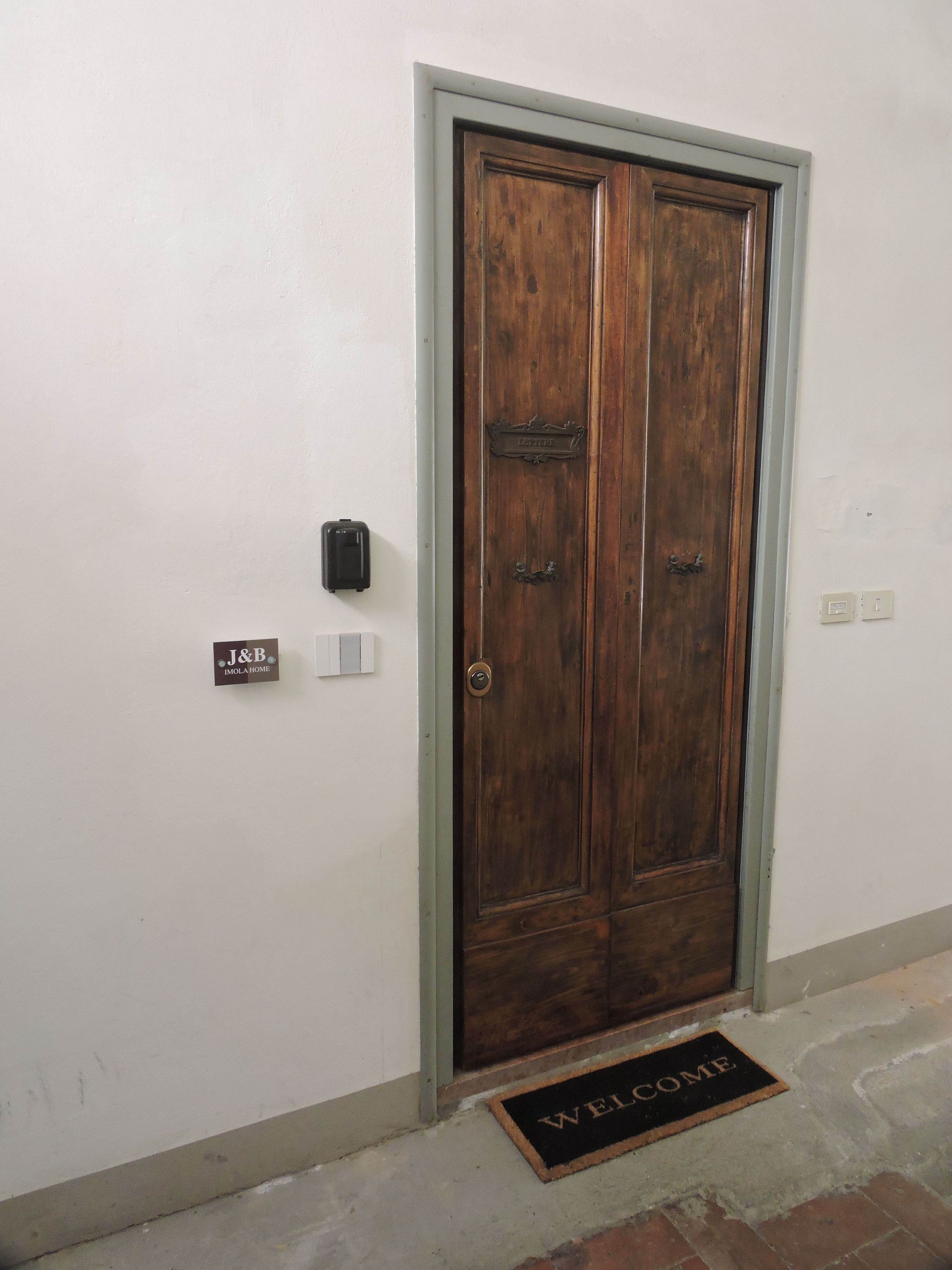 Eine Holztür mit doppelten Flügeln, einer Fußmatte mit der Aufschrift 'WELCOME' und einem Schild, auf dem 'J&B IMOLA HOME' steht.