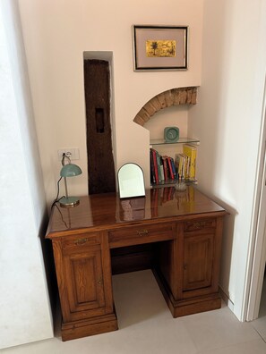 A wooden desk with a glass top, a lamp, a mirror, and a bookshelf with books.