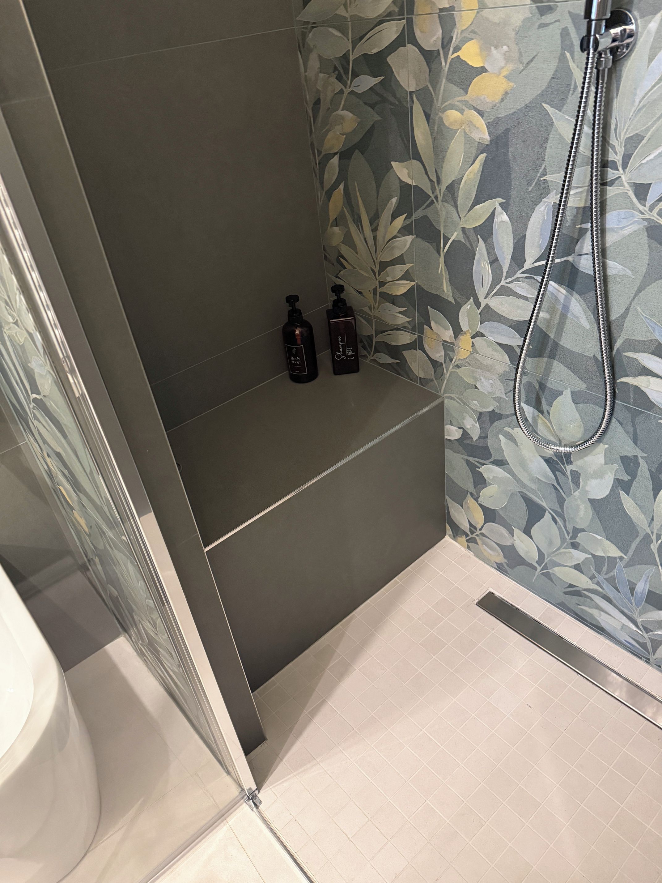 Zona doccia con porta in vetro, soffione e due flaconi di shampoo su un ripiano.
