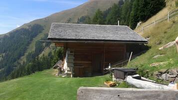 Une cabane en bois, dotée d’un toit en pente, située sur une colline verdoyante, avec une chaîne de montagnes en arrière-plan.