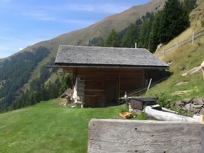 Une cabane en bois, dotée d’un toit en pente, située sur une colline verdoyante, avec une chaîne de montagnes en arrière-plan.
