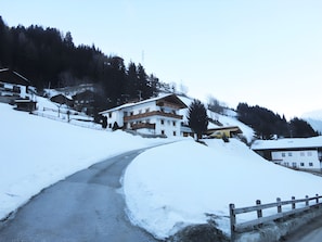 A snow-covered road leading to a multi-story building with a balcony, surrounded by trees and a mountain backdrop. - Apartment '2' with Mountain View, Private Terrace and Wi-Fi (Selva dei Molini)