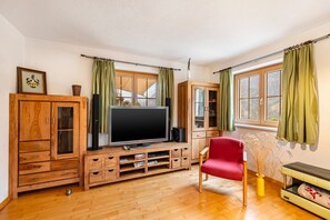 A living room with a wooden entertainment unit, a flat-screen TV, a red chair, and a window with green curtains. - Apartment 'Rackhof' with Mountain View, Private Terrace and Wi-Fi (Oberlienz)