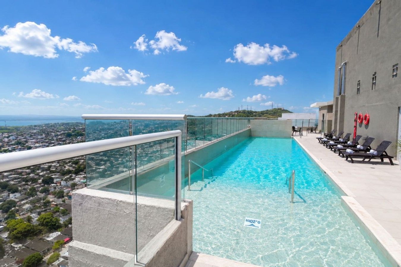A rooftop swimming pool with clear blue water, a glass railing, and a view of the cityscape.