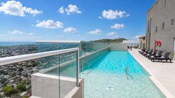 A rooftop swimming pool with clear blue water, a glass railing, and a view of the cityscape.