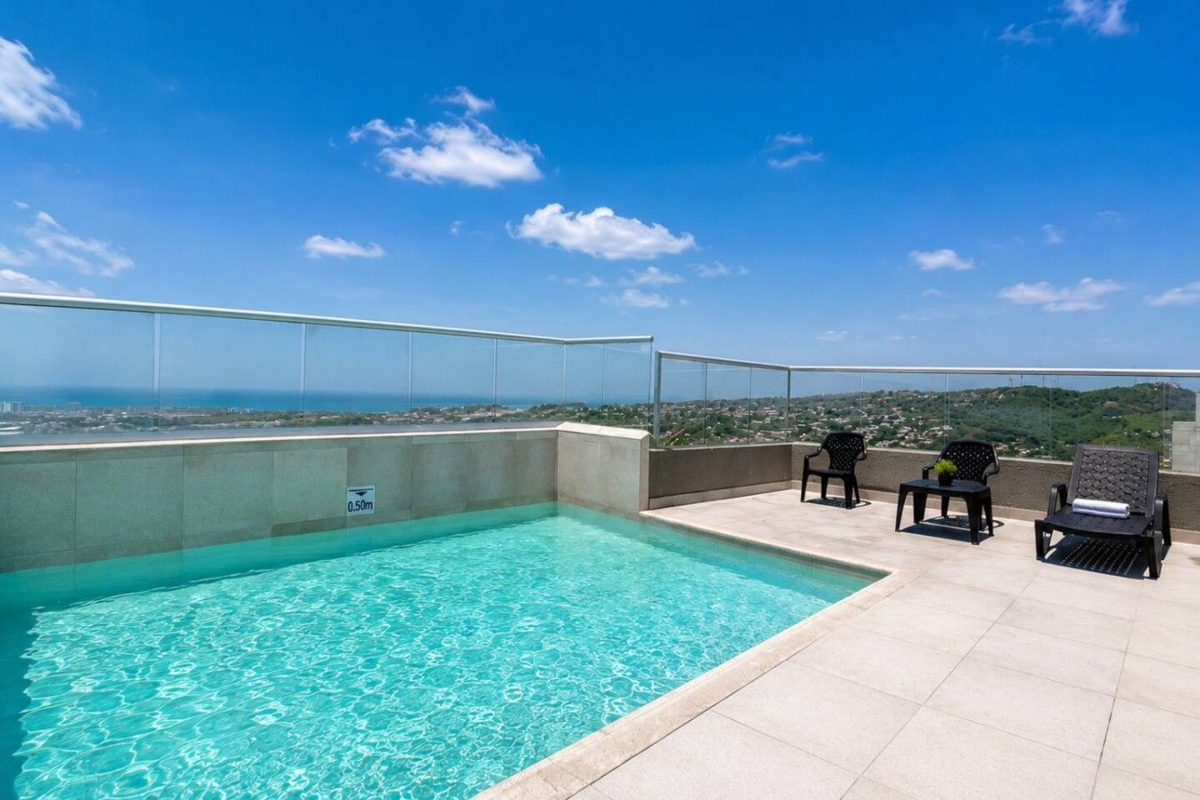 Una piscina en la azotea con agua cristalina y azul, rodeada por una barandilla de vidrio y con vistas a un paisaje urbano.