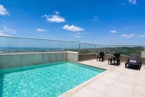 Una piscina en la azotea con agua cristalina y azul, rodeada por una barandilla de vidrio y con vistas a un paisaje urbano.