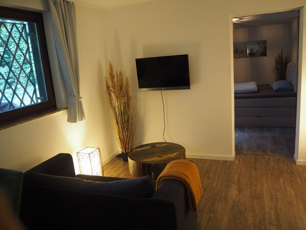 Una sala de estar con sofá, un televisor de pantalla plana montado en la pared, una mesita auxiliar con una planta y una ventana con cortinas.