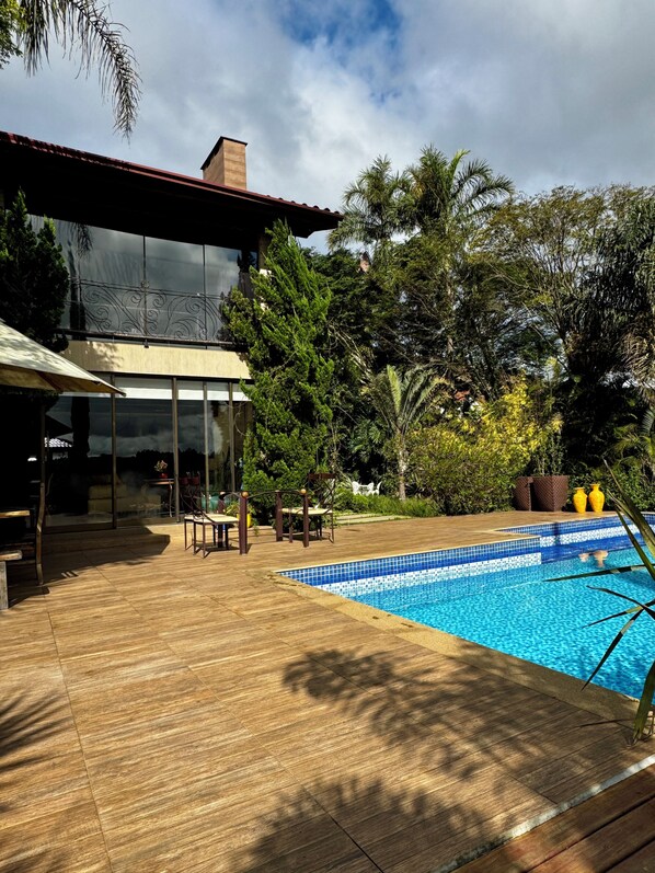 A modern outdoor pool area with a wooden deck, a glass-enclosed patio, and a variety of trees and plants.