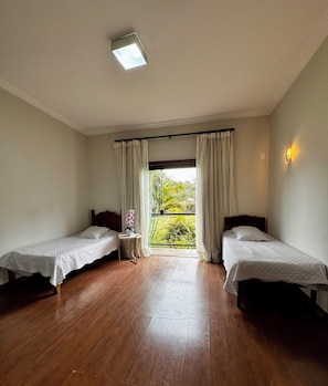 A hotel room with two single beds, a small table with a vase of flowers, and a view of greenery through a window.