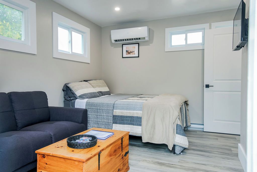 A compact bedroom with a bed, a sofa, a wooden chest, and an air conditioning unit.