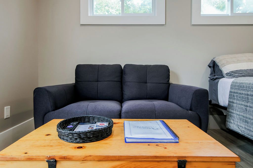 Une pièce avec un canapé gris foncé, une table en bois sur laquelle se trouvent un panier et un carnet, et un lit recouvert d’une couverture à rayures.