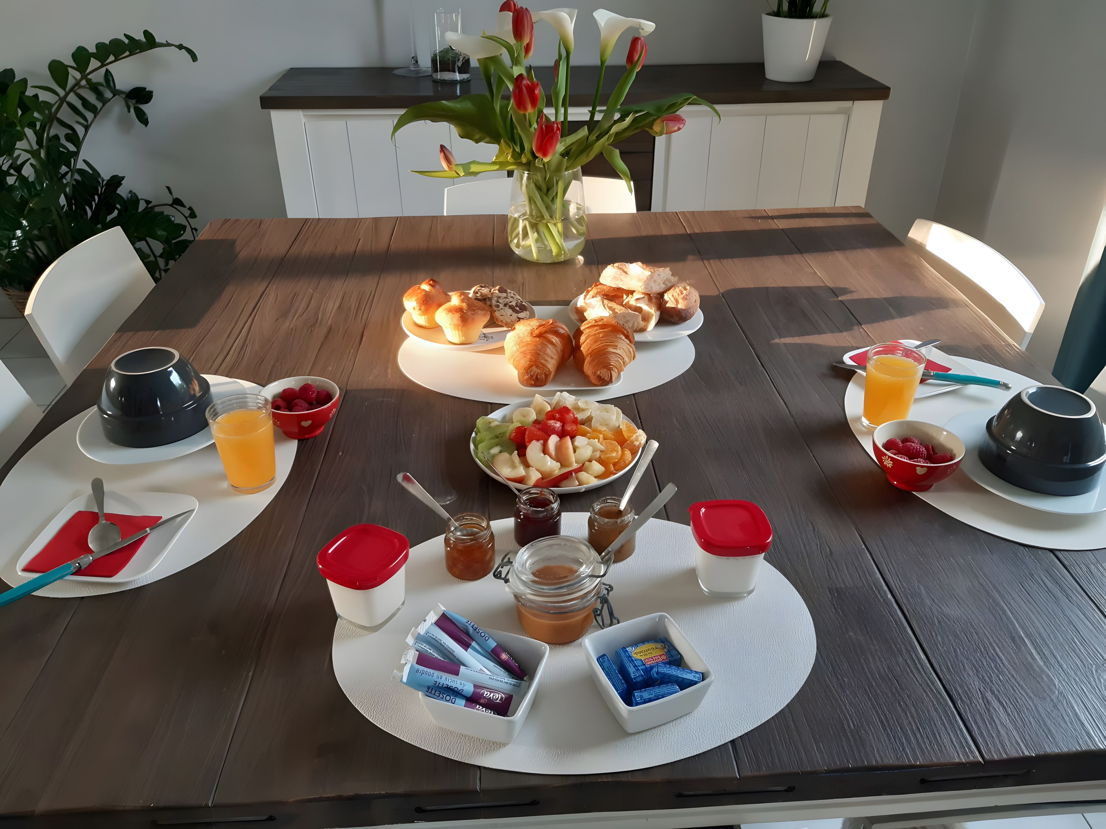 Une table de petit-déjeuner dressée avec des croissants, des muffins, des fruits et des condiments.
