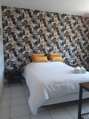 A hotel room with a patterned wallpaper, a bed with white bedding and two yellow pillows, a bedside table with a lamp, and a rolled towel on the bed. - Bed and Breakfast 'Escale Sabolienne' with Shared Garden and Wi-Fi (Sablé-sur-Sarthe)