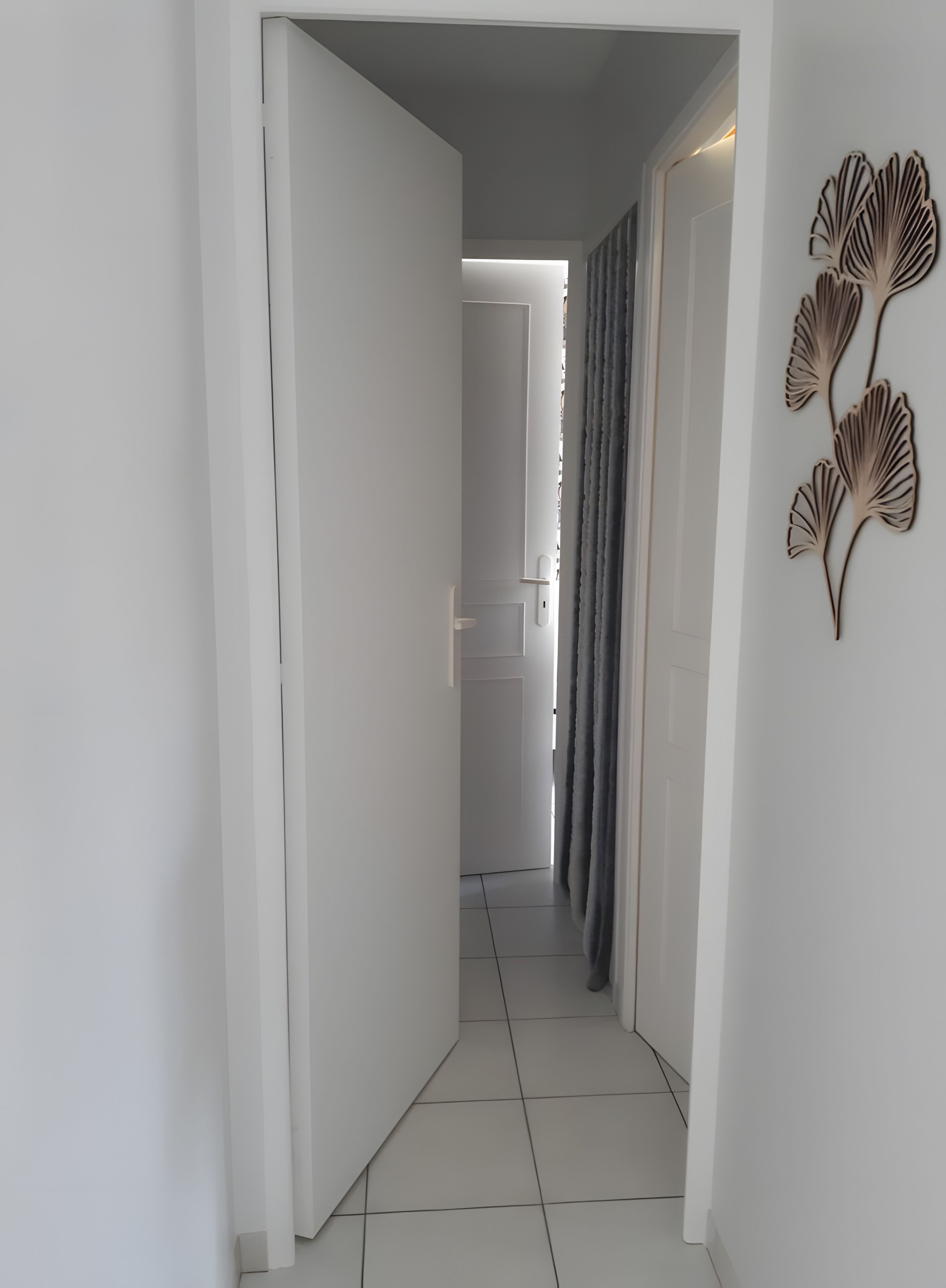 Un corridoio con pareti bianche, pavimento piastrellato e una porta che dà su un'altra stanza. Sulla destra si nota un elemento decorativo a parete.