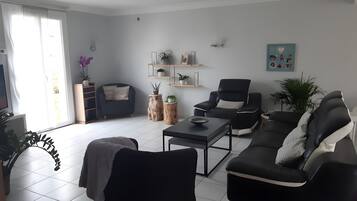 A modern living room with a sofa, armchairs, a coffee table, and a TV. There are plants and shelves with decorative items.