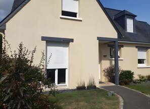 A two-story house with a beige exterior, a covered porch, and a small garden area with plants and a pathway. - Bed and Breakfast 'Escale Sabolienne' with Shared Garden and Wi-Fi (Sablé-sur-Sarthe)