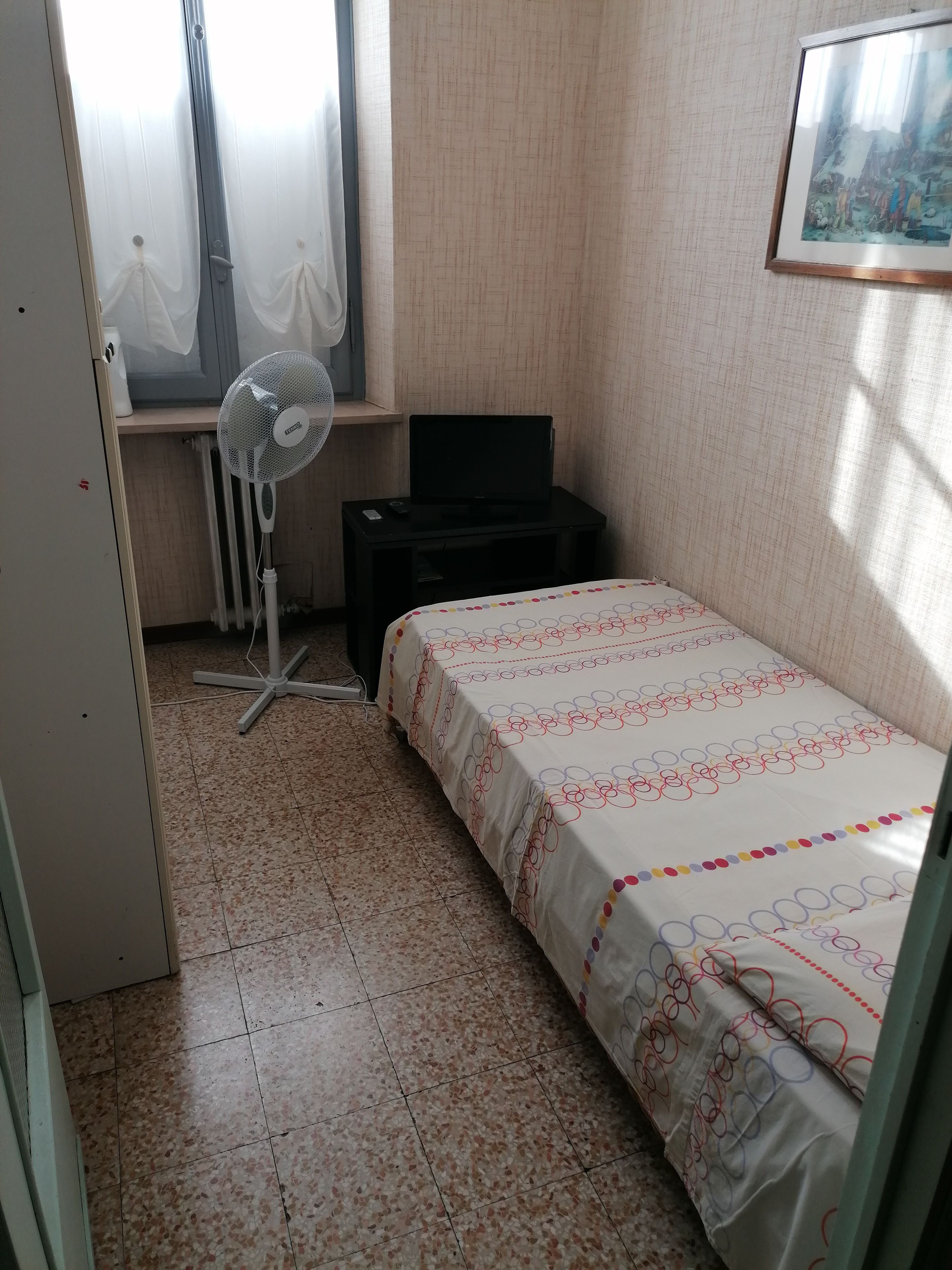 Ein kleines Schlafzimmer mit einem Bett, einem Standventilator, einem schwarzen Fernseher auf einem kleinen Tisch und einem Fenster mit durchsichtigen Vorhängen.