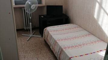 Ein kleines Schlafzimmer mit einem Bett, einem Standventilator, einem schwarzen Fernseher auf einem kleinen Tisch und einem Fenster mit durchsichtigen Vorhängen.
