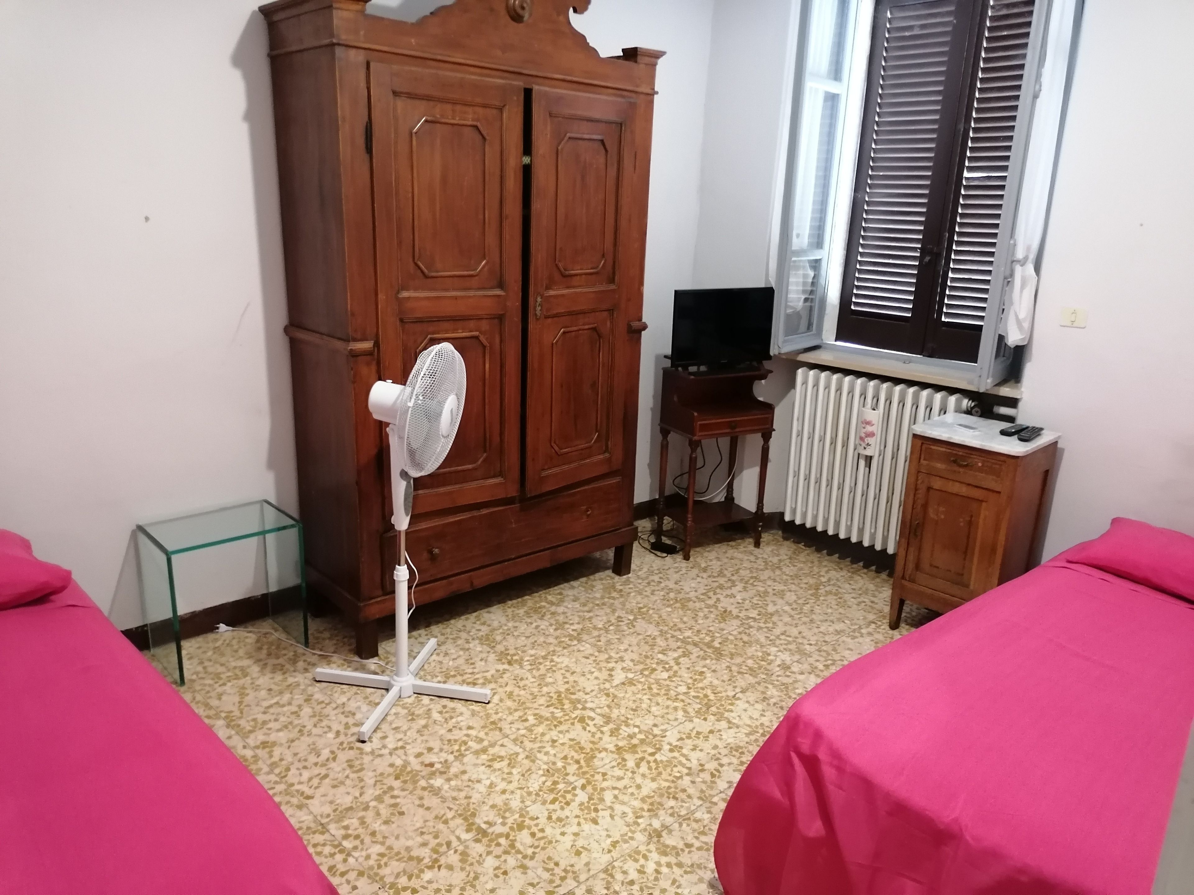 Un dormitorio con un armario de madera, un ventilador de pie, un radiador, un televisor y dos camas con cubrecamas rosas.