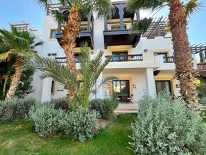 Ein mehrstöckiges Gebäude mit Balkonen, ein Garten mit verschiedenen Sträuchern und einer Terrasse mit Gartenmöbeln.