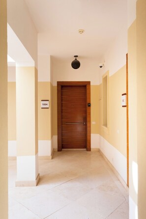 A hallway with a wooden door, a ceiling light, and a wall-mounted security camera. - VESTA - Luxury APT - 1BR - Ancient sands (El Gouna)