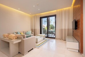 A modern living room with a sofa, a coffee table, and a TV. There is a sliding glass door leading to an outdoor area with a table and chairs. - VESTA - Luxury APT - 1BR - Ancient sands (El Gouna)