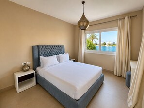 Ein Schlafzimmer mit einem großen Bett, einem Nachttisch, einer Nachttischlampe, einem Fenster mit Vorhängen und Blick aufs Meer.