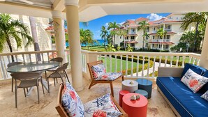 A balcony with a dining table, chairs, and a sofa with cushions, overlooking a resort with palm trees and a building with balconies. - Ocean View Retreat • Steps to Private Beach • Free Weekly Maid Service (Sosúa)