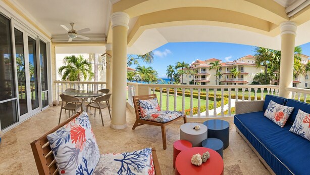 A balcony with a sofa, chairs, and a table, overlooking a resort with multiple buildings and palm trees. - Ocean View Retreat • Steps to Private Beach • Free Weekly Maid Service (Sosúa)