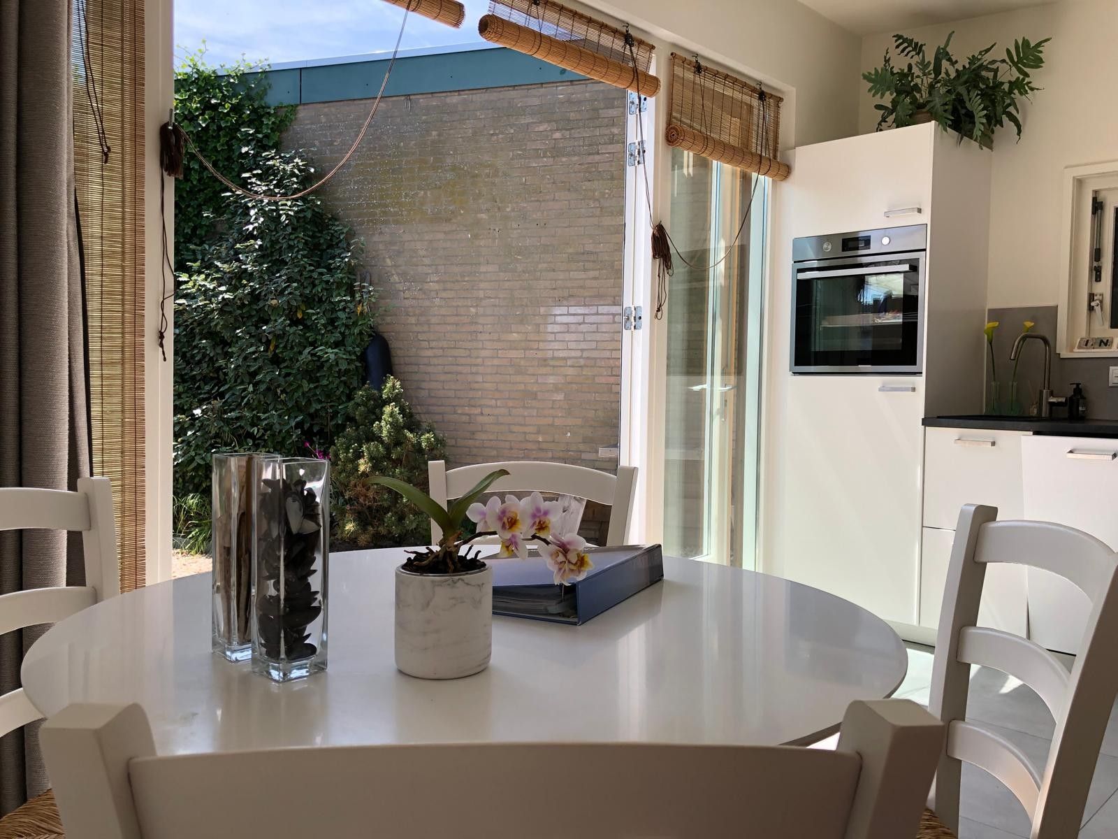 A modern kitchen with a built-in oven, a dining table with chairs, and a view of a brick wall and greenery outside.