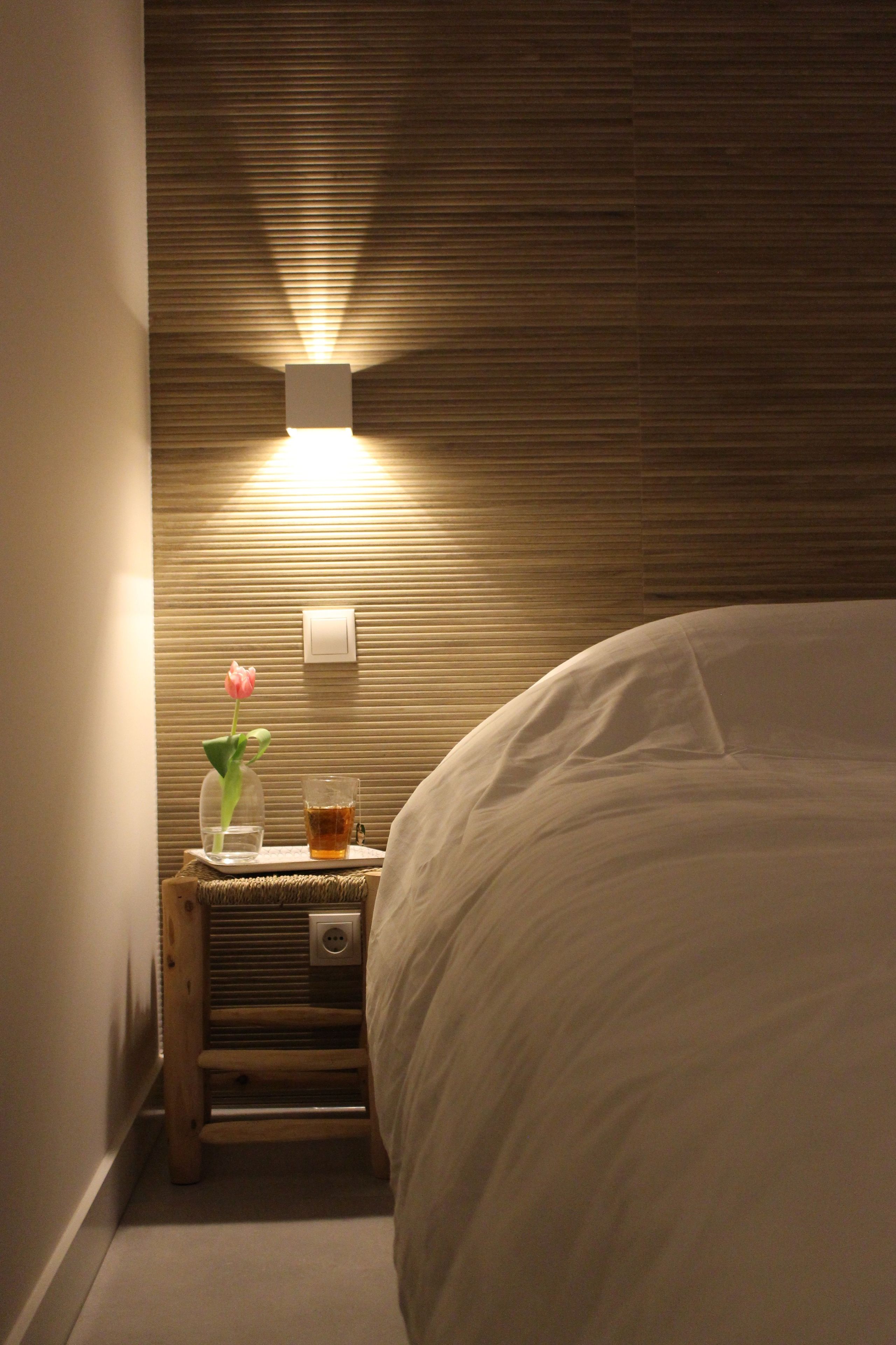 Hotellihuone, jossa on sänky, yöpöytä maljakko täynnä kukkia, lasi vettä ja seinään kiinnitetty valaisin.