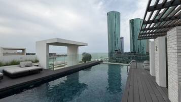 Un espace piscine sur le toit, doté d’un design moderne, comprenant une balustrade en verre, une terrasse en bois et un coin salon avec des coussins.