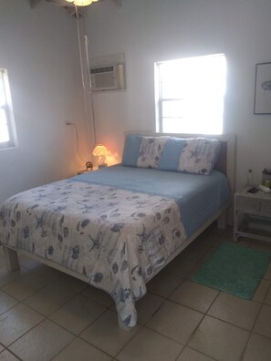 Ein Schlafzimmer mit einem Bett, einem Nachttisch, einer Lampe, einem Fenster und einer Wand-Klimaanlage.