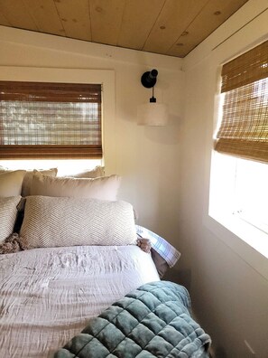 Une chambre à coucher avec un lit, des oreillers, une couette, un luminaire suspendu et une fenêtre avec des stores.