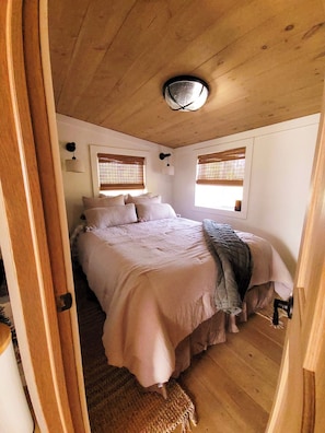 Un dormitorio con techo de madera, una cama con ropa de cama blanca, dos ventanas con persianas y una puerta.