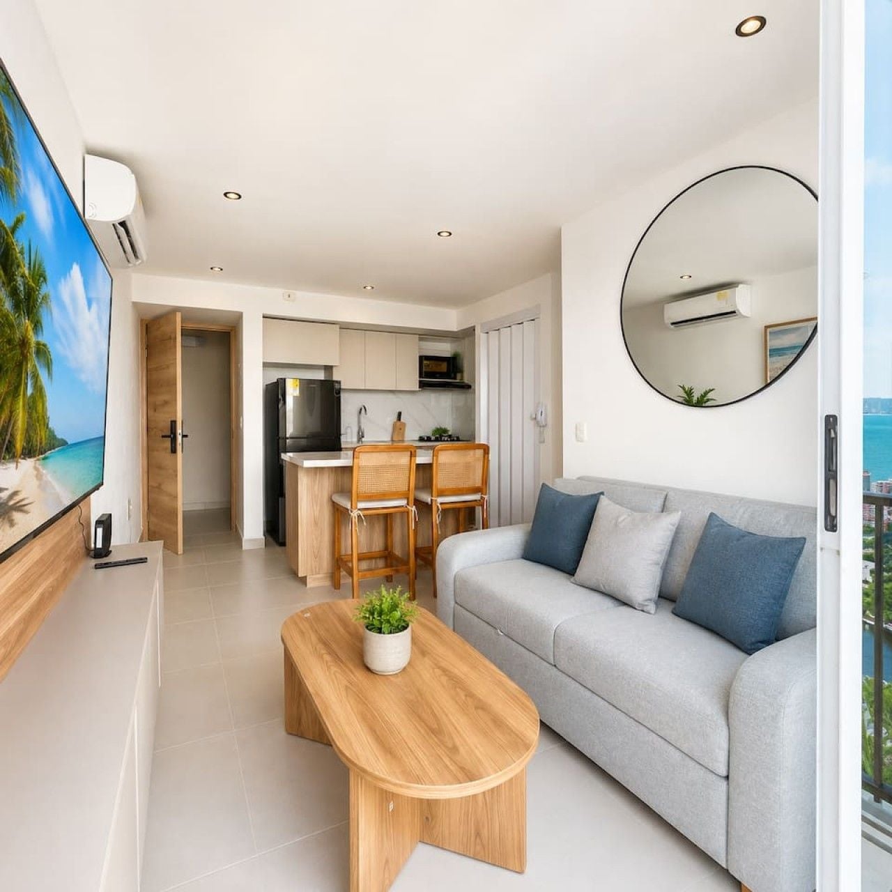 A modern living room with a sofa, a coffee table, and a TV. There is a kitchen area with a dining set and an island.