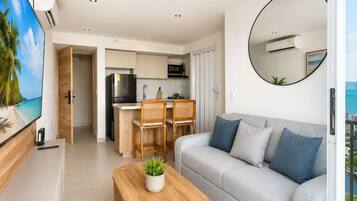 A modern living room with a sofa, a coffee table, and a TV. There is a kitchen area with a dining set and an island.