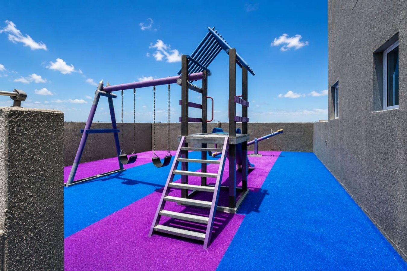 Zona infantil con columpios, un tobogán y una escalera sobre un suelo azul y morado.