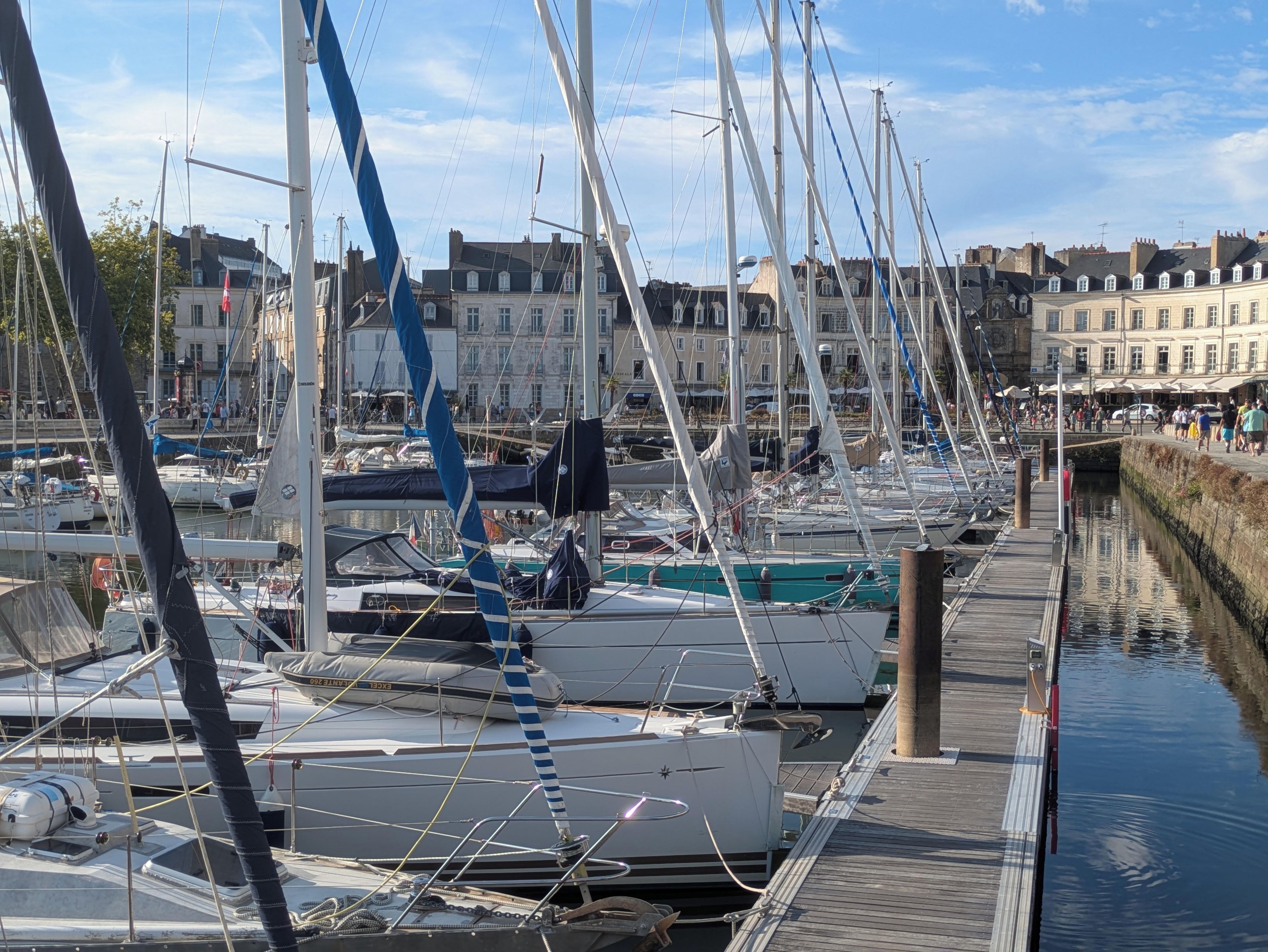 Un port de plaisance où sont amarrés de nombreux bateaux, une passerelle en bois et des bâtiments en arrière-plan.