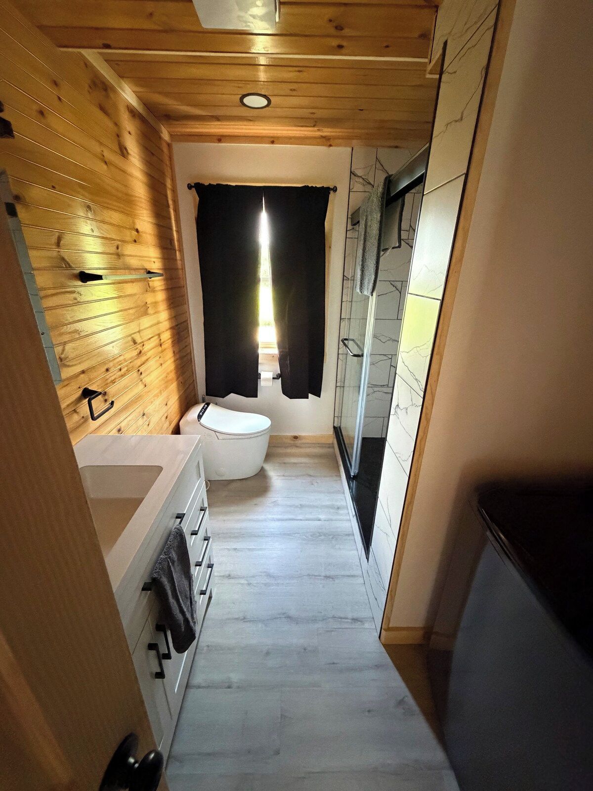 A narrow bathroom with a wooden ceiling, a white toilet, a sink with a towel, and a glass shower enclosure.