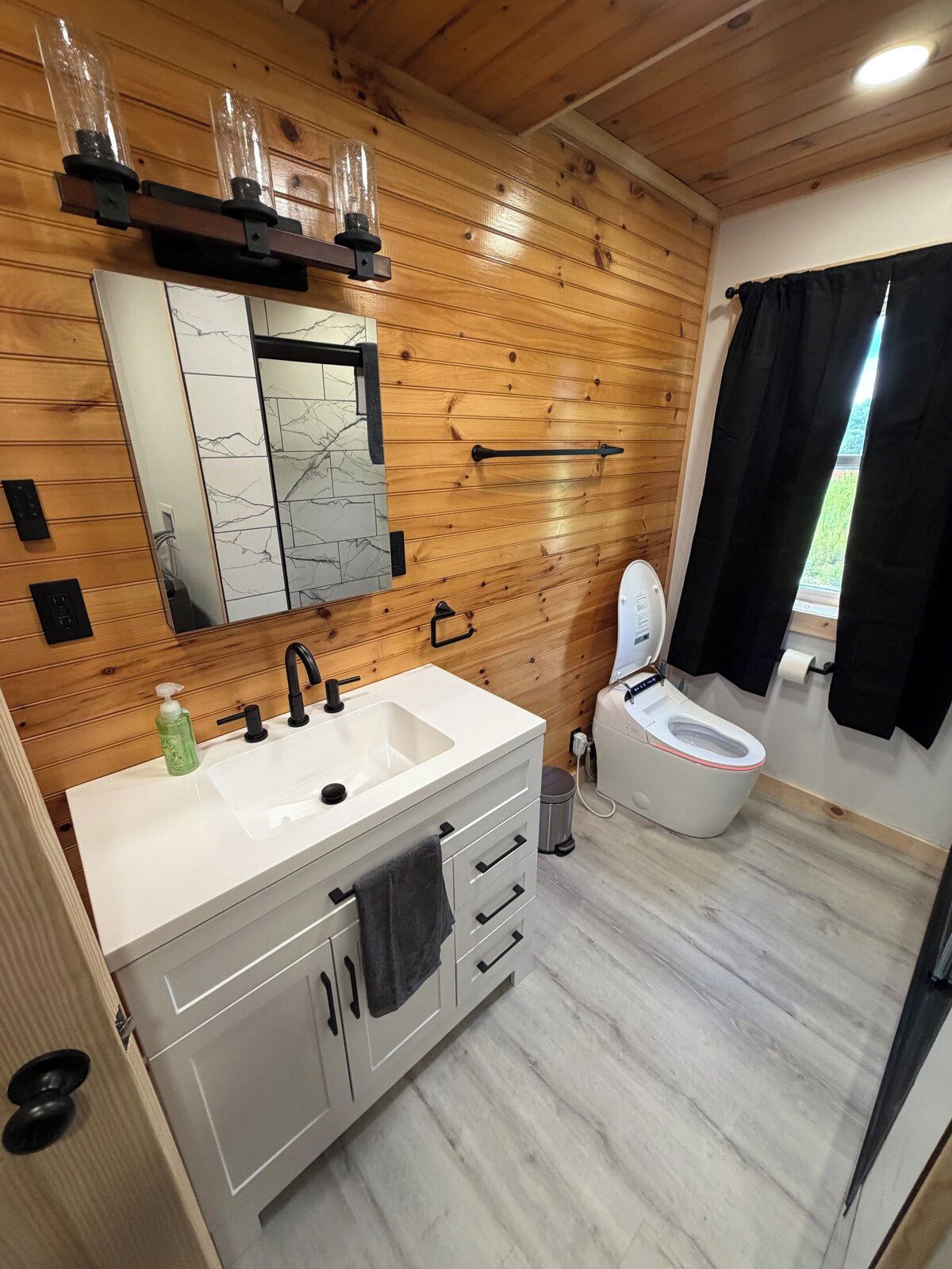 A modern bathroom with a wooden wall, a white sink with a mirror, a toilet, and a window with a view.