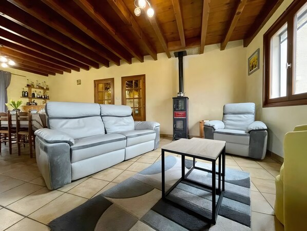 A living room with a sofa, armchairs, a table, a fireplace, and a dining area with chairs and a shelf of bottles.