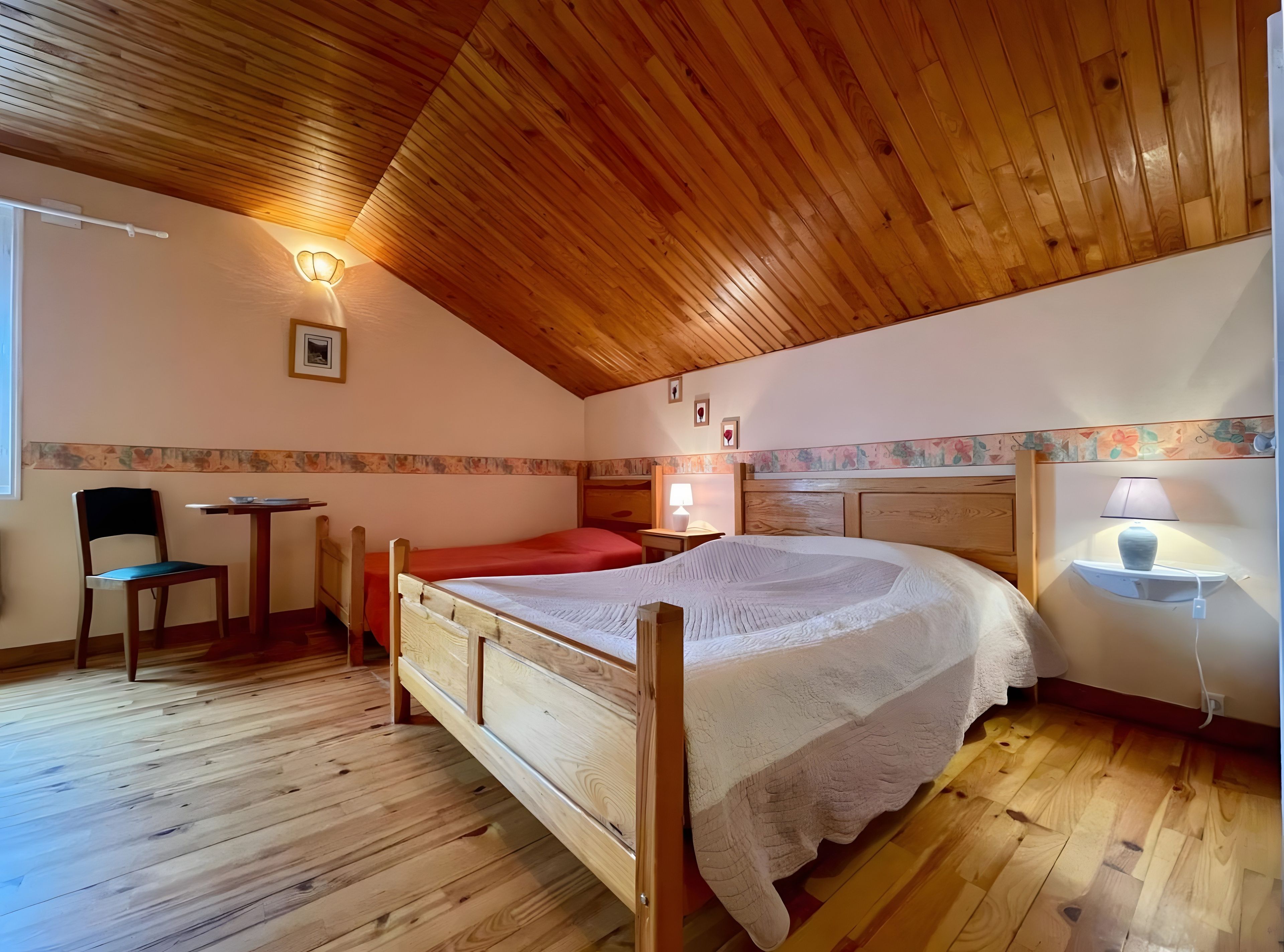 A bedroom with a wooden ceiling, a single bed, a chair, a small table, and a nightstand with a lamp.