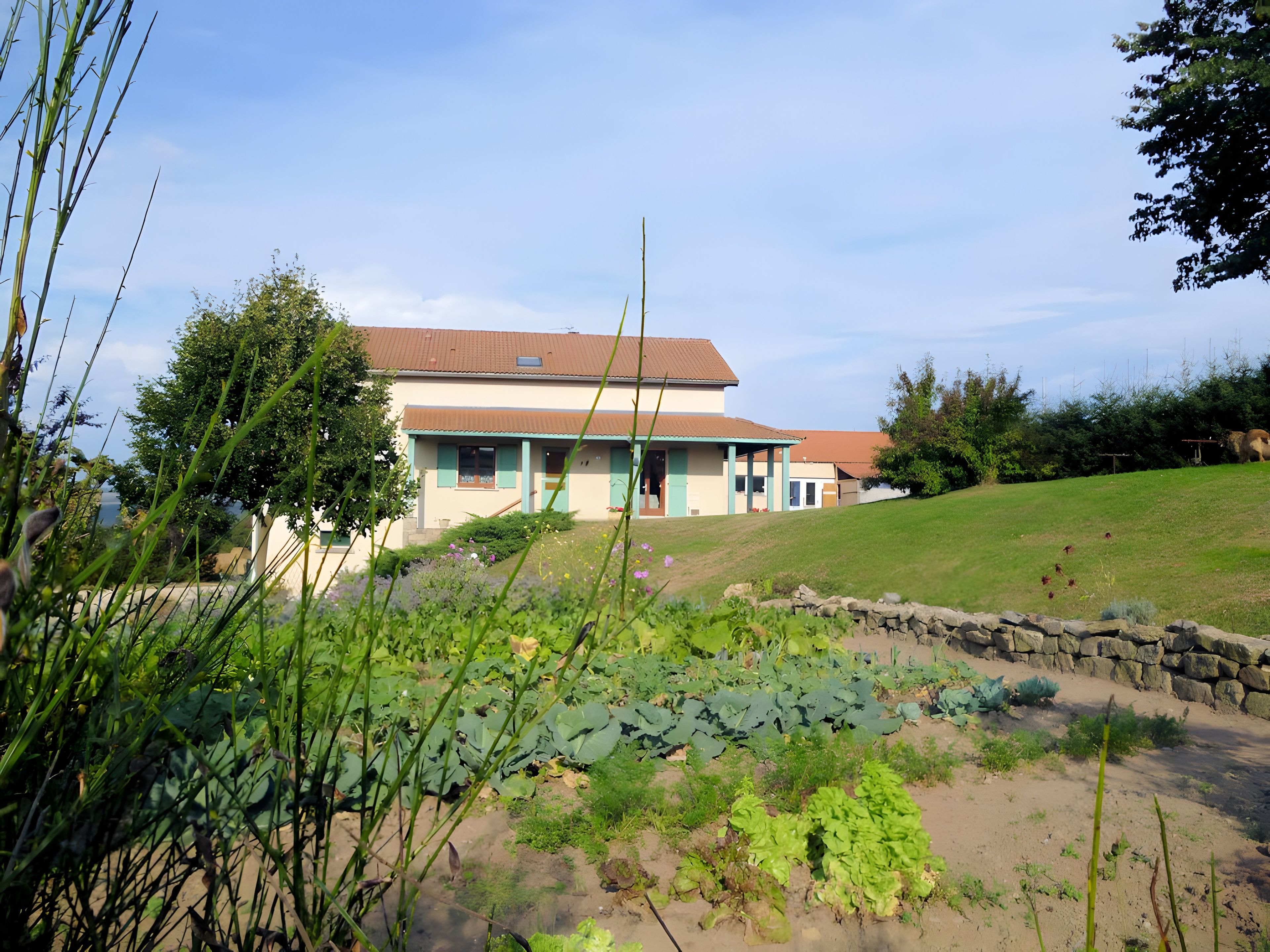 Ein zweistöckiges Haus mit geneigtem Dach, umgeben von einem Garten mit verschiedenen Pflanzen und einer Steinmauer.