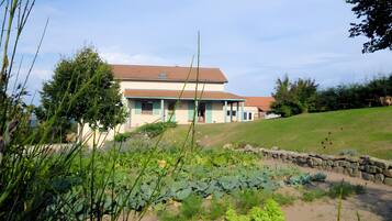 Ein zweistöckiges Haus mit geneigtem Dach, umgeben von einem Garten mit verschiedenen Pflanzen und einer Steinmauer.