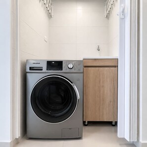 A compact laundry room with a front-loading washing machine, a wooden cabinet, and a white tiled wall. - Royal Palm | Old Town Views, Marbella Beach & Pool (Cartagena)