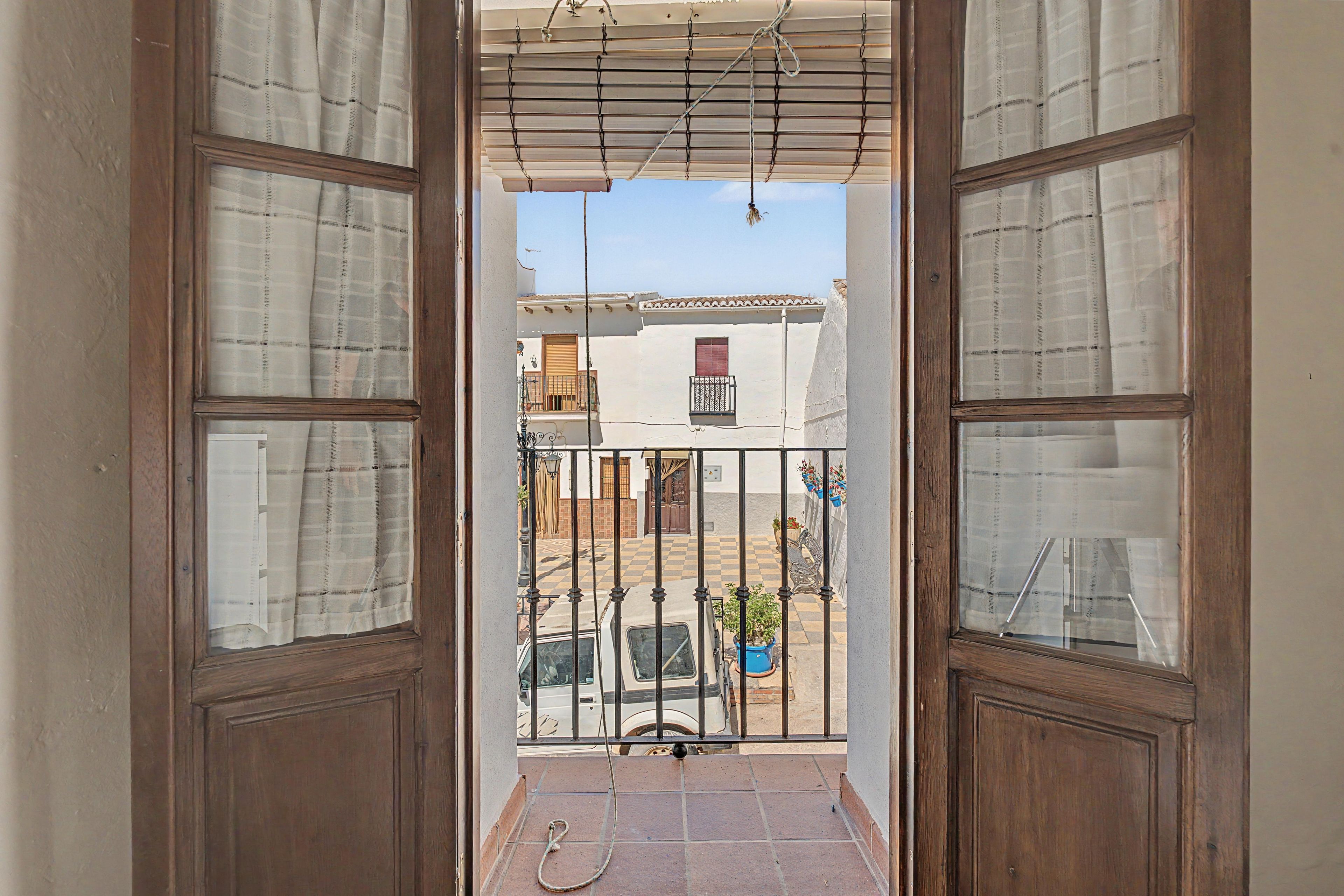 Une vue à travers une porte en bois, avec un sol à carreaux et un portail en métal donnant sur une cour où se trouve une voiture garée et un pot de fleurs.