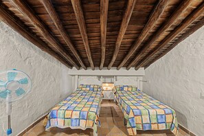 Una habitación con techo de madera, dos camas con ropa de cama colorida, una pared blanca y un ventilador.
